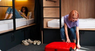 Girls packing red suitcase in hostel room with bunk beds.